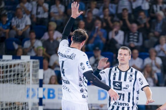 Zwei männliche Handballspieler in schwarz-weiß gestreiften Trikots stehen auf einem Hallenplatz. Ein Spieler hebt die Hand zum Abklatschen, während der andere ihn anschaut. Im unscharfen Hintergrund sind Zuschauer zu sehen.