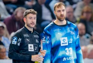 Zwei männliche Handballspieler stehen während eines Spiels Seite an Seite. Der eine trägt ein schwarzes Trikot und spricht, während er eine Wasserflasche in der Hand hält; der andere trägt ein blaues Trikot mit der Nummer 33. Im Hintergrund ist eine unscharfe Menschenmenge zu sehen.