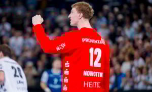 Ein Handballspieler, der ein leuchtend rotes Trikot mit der Nummer 12 und dem Namen Nowotny trägt, jubelt mit erhobener Faust in einer Halle, mit einer unscharfen Menge und anderen Spielern im Hintergrund.