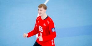 Ein junger männlicher Handballspieler, der ein rotes Trikot mit der Nummer 12 trägt, lächelt und ballt seine Fäuste zum Jubel auf einem blauen Hallenplatz.