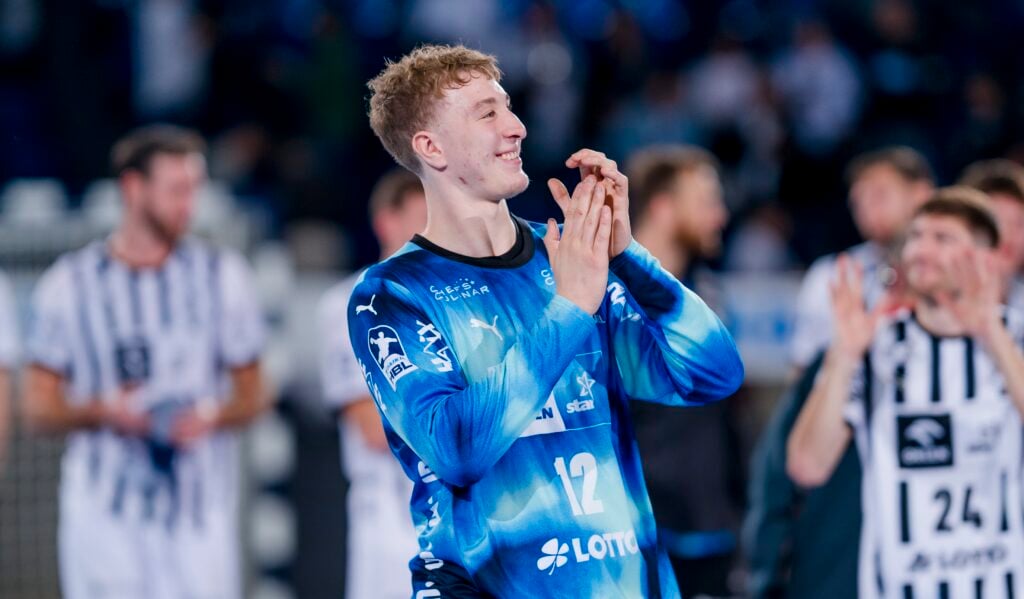 Ein männlicher Handballspieler in einem blauen Trikot mit der Nummer 12 klatscht und lächelt, während er auf einem Spielfeld steht. Andere Spieler in schwarz-weiß gestreiften Trikots sind im Hintergrund zu sehen. Die Szene wirkt feierlich.
