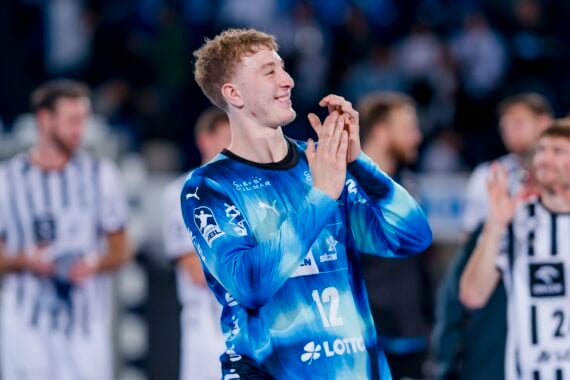 Ein männlicher Handballspieler in einem blauen Trikot mit der Nummer 12 klatscht und lächelt, während er auf einem Spielfeld steht. Andere Spieler in schwarz-weiß gestreiften Trikots sind im Hintergrund zu sehen. Die Szene wirkt feierlich.