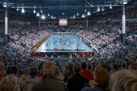 Eine überfüllte Halle mit Zuschauern, die ein Handballspiel auf einem gut beleuchteten Platz verfolgen, umgeben von Sitzreihen und einer großen Anzeigetafel, die über der Mitte des Platzes hängt.
