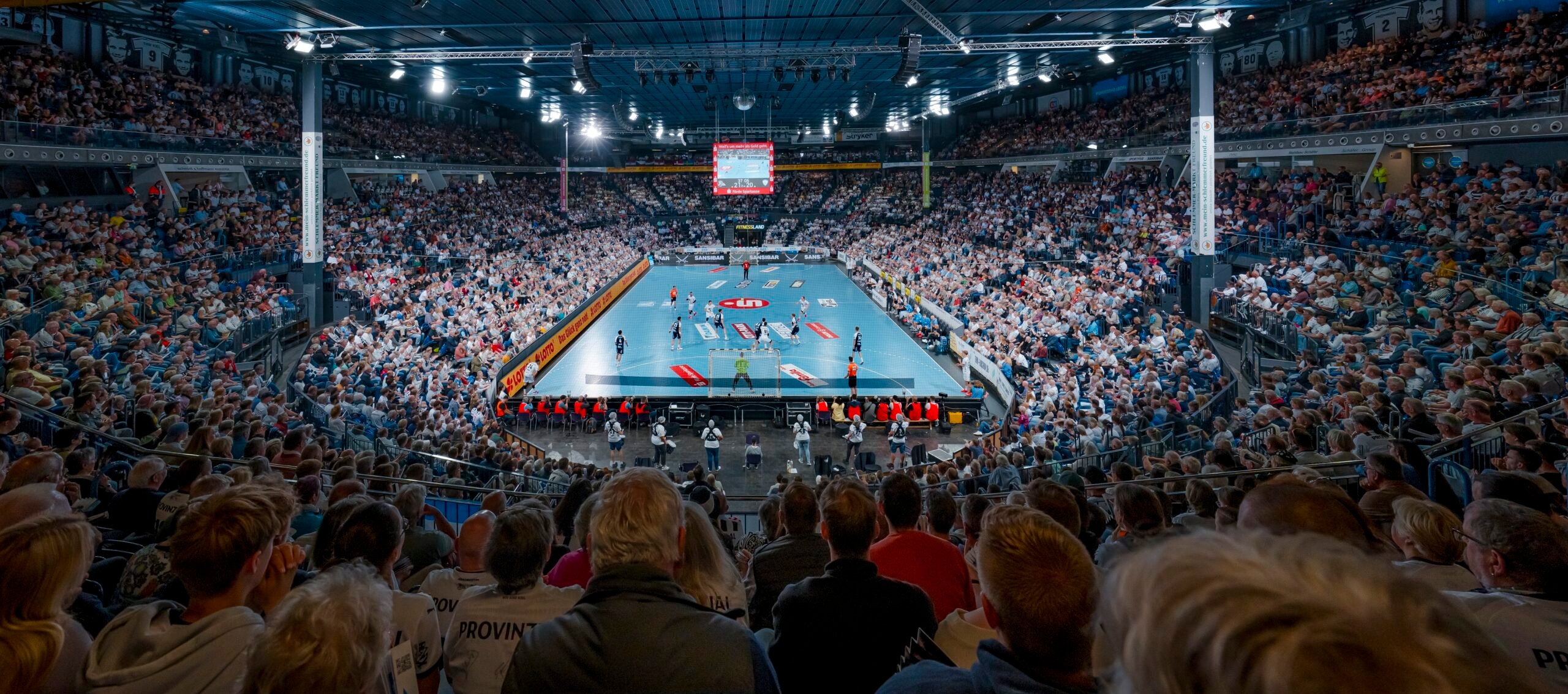 Eine überfüllte Halle mit Zuschauern, die ein Handballspiel auf einem gut beleuchteten Platz verfolgen, umgeben von Sitzreihen und einer großen Anzeigetafel, die über der Mitte des Platzes hängt.