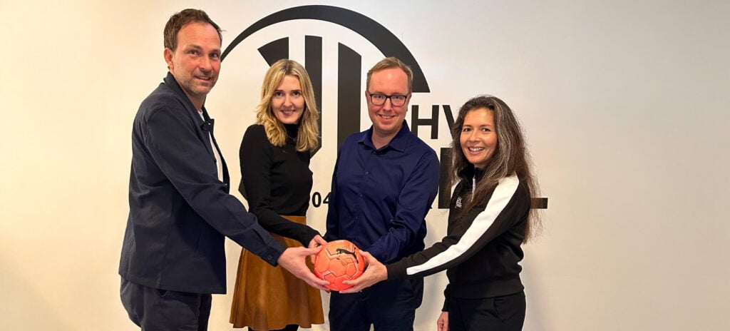 Vier Personen stehen vor einer weißen Wand mit einem schwarzen Logo, lächeln und halten einen orangefarbenen Handball zusammen. Zwei Männer stehen links und in der Mitte, zwei Frauen stehen rechts und in der Mitte.
