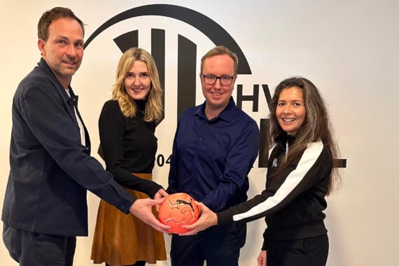 Vier Personen stehen vor einer weißen Wand mit einem schwarzen Logo, lächeln und halten einen orangefarbenen Handball zusammen. Zwei Männer stehen links und in der Mitte, zwei Frauen stehen rechts und in der Mitte.