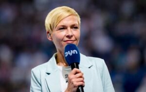 Eine Frau mit kurzem blondem Haar hält ein blaues Mikrofon mit der Aufschrift dyn. Sie trägt einen hellblauen Blazer und steht vor einer unscharfen Menge, die den Eindruck erweckt, als würde sie auf einer Veranstaltung sprechen oder moderieren.