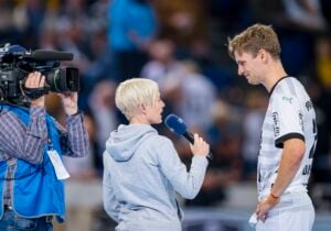 Eine Reporterin mit kurzen blonden Haaren interviewt einen männlichen Sportler in einem weißen Trikot, während ein Kameramann sie filmt; sie stehen auf einem Sportplatz mit einer verschwommenen Menschenmenge im Hintergrund.
