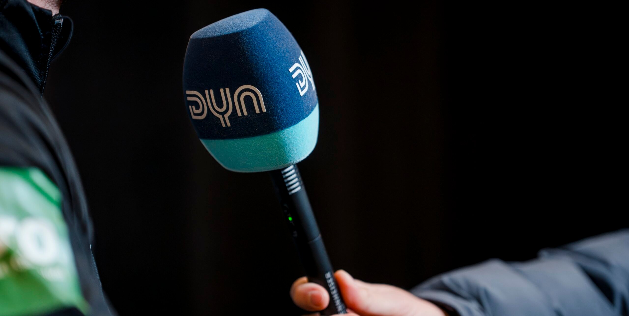 Eine Person hält ein blaues und blaugrünes Mikrofon der Marke DYN in der Nähe einer Person, die eine dunkle Jacke trägt und möglicherweise ein Interview führt. Der Hintergrund ist dunkel und unscharf.