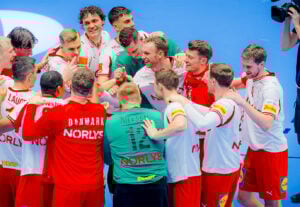 Eine Gruppe von männlichen Handballspielern in weißen und roten Trikots steht auf einem blauen Spielfeld zusammen, feiert und lächelt. Der Spieler in der Mitte trägt ein grünes Trikot mit der Nielsen 12 auf dem Rücken.