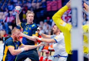 Ein Handballspieler in einem blau-gelben Trikot springt zum Schuss, während er von Gegnern in weißen Trikots verteidigt wird. Im Hintergrund sind andere Spieler und eine Menschenmenge zu sehen. Die Szene ist actionreich und intensiv.