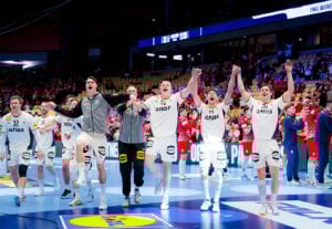 Eine Männer-Handballmannschaft in weißen Trikots feiert auf einem blauen Hallenplatz, hält sich an den Händen und springt vor Freude, während jubelnde Fans in Rot den Hintergrund füllen.