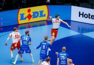 Ein männlicher Handballspieler in einem weiß-roten Trikot springt, um den Ball zu schießen, während blau gekleidete Verteidiger versuchen, ihn während eines Spiels zu blockieren. Im Hintergrund ist ein großes Lidl-Logo zu sehen.