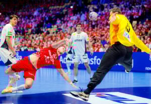 Ein Handballspieler in einem roten Trikot springt und wirft den Ball in Richtung Tor, während sich ein Torwart in einem gelben Trikot streckt, um ihn zu blockieren; im Hintergrund sind andere Spieler und eine Menschenmenge zu sehen.