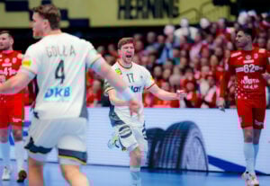 Ein Handballspieler in einem weißen Trikot mit der Nummer 17 feiert ausgelassen auf dem Spielfeld. Im Hintergrund sind andere Spieler in weißen und roten Trikots sowie eine Menschenmenge zu sehen.