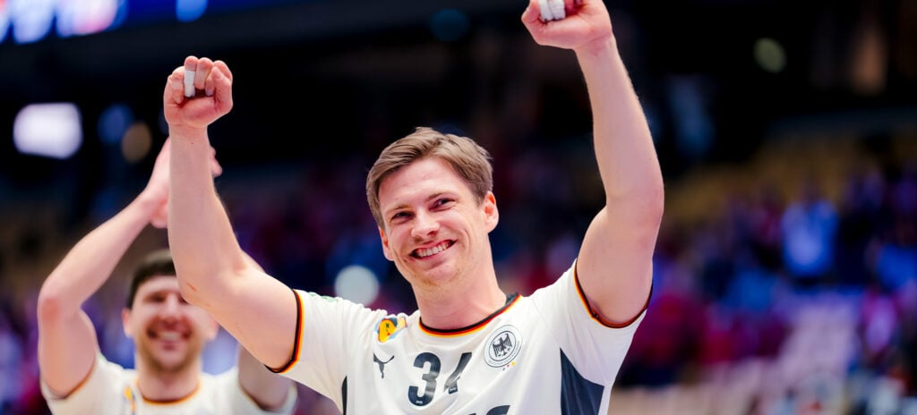 260131_Titel_EM-Update_Dahmke Ein lächelnder männlicher Sportler in einem weißen Sporttrikot mit der Nummer 31 hebt beide Arme zum Jubel, während die Menge im Hintergrund verschwimmt. Ein anderer Mannschaftskamerad ist zu sehen, der ebenfalls jubelt.