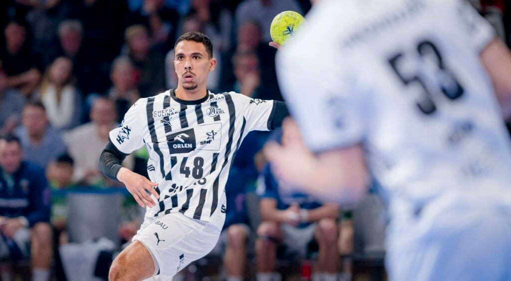 HANDBALL 2025/26: Füchse Berlin - THW Kiel Ein männlicher Handballspieler in einem schwarz-weiß gestreiften Trikot mit der Nummer 48 bereitet sich darauf vor, während eines Spiels einen gelben Ball zu werfen. Ein anderer Spieler in einem weißen Trikot mit der Nummer 53 ist teilweise im Vordergrund zu sehen. Im Hintergrund sind Zuschauer zu sehen.