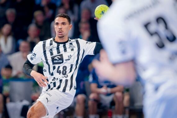 Ein männlicher Handballspieler in einem schwarz-weiß gestreiften Trikot mit der Nummer 48 bereitet sich darauf vor, während eines Spiels einen gelben Ball zu werfen. Ein anderer Spieler in einem weißen Trikot mit der Nummer 53 ist teilweise im Vordergrund zu sehen. Im Hintergrund sind Zuschauer zu sehen.