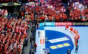 Eine Handballmannschaft in roten Trikots steht auf dem Spielfeld vor einer jubelnden, rot gekleideten Menge in einer mit Zuschauern gefüllten Arena. Rund um das Spielfeld sind Banner und Werbung zu sehen.
