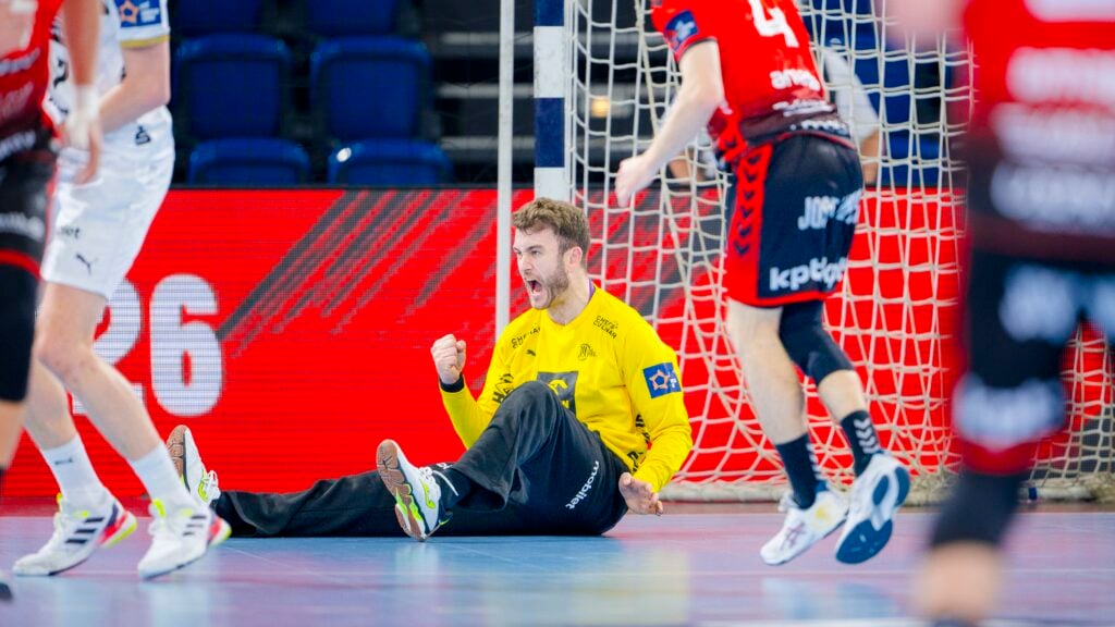 Ein Handballtorwart im gelben Trikot sitzt mit geballter Faust auf dem Boden und feiert, während Spieler in roten und weißen Trikots während des THW Kiel Handball-Derbys in der EHF European League Saison 2025/2026 in der Nähe laufen.