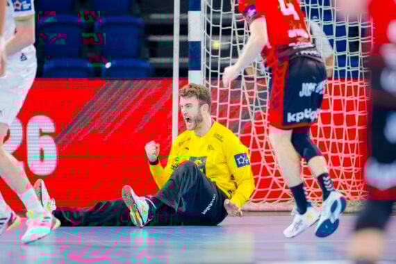 Ein Handballtorwart im gelben Trikot sitzt mit geballter Faust auf dem Boden und feiert, während Spieler in roten und weißen Trikots während des THW Kiel Handball-Derbys in der EHF European League Saison 2025/2026 in der Nähe laufen.