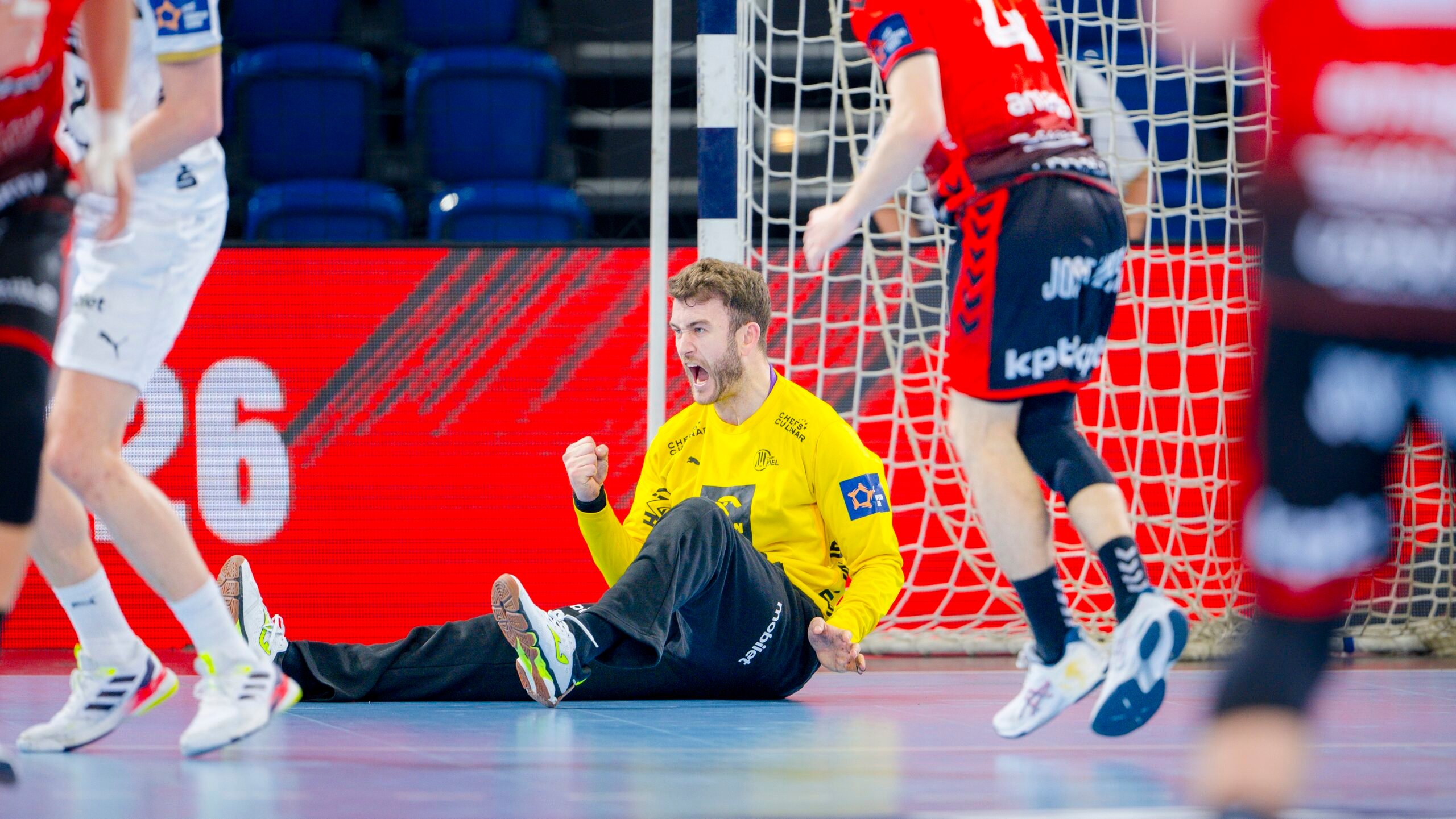 Ein Handballtorwart im gelben Trikot sitzt mit geballter Faust auf dem Boden und feiert, während Spieler in roten und weißen Trikots während des THW Kiel Handball-Derbys in der EHF European League Saison 2025/2026 in der Nähe laufen.