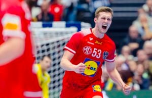 Ein männlicher Handballspieler in einem roten Trikot mit der Nummer 53 feiert leidenschaftlich auf dem Spielfeld mit einer geballten Faust, den Mund zu einem Schrei geöffnet. Im Hintergrund sind unscharfe Zuschauer und andere Spieler zu sehen.