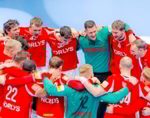 Eine Gruppe männlicher Athleten in roten und grünen Trikots steht vor einem Spiel zusammen, legt die Arme umeinander, lächelt und jubelt energisch.