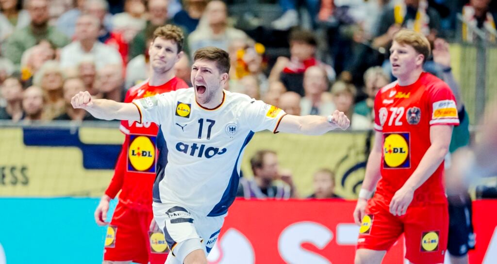 Ein männlicher Handballspieler in einem weißen Trikot steht während eines Spiels mit erhobenen Armen und geöffnetem Mund vor zwei gegnerischen Spielern in roten Trikots. Im Hintergrund ist eine jubelnde Menge zu sehen.