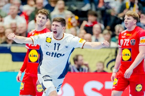 Ein männlicher Handballspieler in einem weißen Trikot steht während eines Spiels mit erhobenen Armen und geöffnetem Mund vor zwei gegnerischen Spielern in roten Trikots. Im Hintergrund ist eine jubelnde Menge zu sehen.