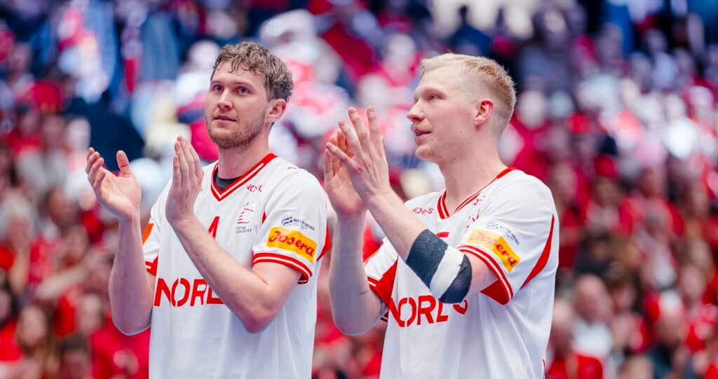 Zwei männliche Athleten in weißen norwegischen Handballtrikots stehen nebeneinander und klatschen, mit einer verschwommenen Menge in Rot hinter ihnen. Beide blicken während einer Sportveranstaltung mit konzentrierter Miene nach vorne.