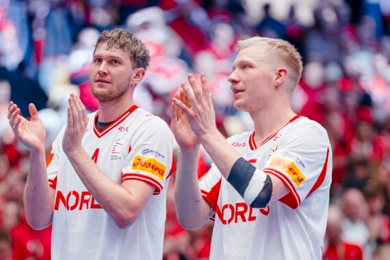 Zwei männliche Athleten in weißen norwegischen Handballtrikots stehen nebeneinander und klatschen, mit einer verschwommenen Menge in Rot hinter ihnen. Beide blicken während einer Sportveranstaltung mit konzentrierter Miene nach vorne.