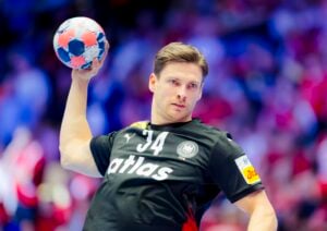 Ein männlicher Handballspieler in einem schwarzen Trikot mit der Nummer 34 bereitet sich darauf vor, einen Ball während eines Spiels zu werfen. Der Hintergrund ist unscharf und zeigt eine Menschenmenge mit roten Farben.