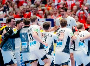 Eine Gruppe männlicher Handballspieler in weißen und grauen Trikots feiert ausgelassen auf dem Spielfeld, wobei ein Spieler vor Begeisterung springt. Eine jubelnde, rot gekleidete Menge füllt den Hintergrund.