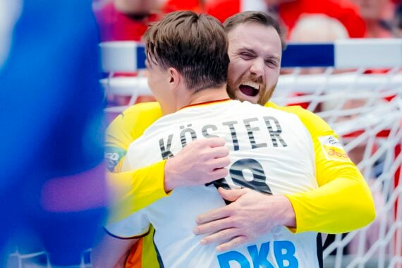 Zwei männliche Handballspieler in weißen Trikots umarmen sich und feiern fröhlich auf dem Spielfeld vor einem Tornetz, wobei der Name Köster" und die Nummer 19 auf dem Rücken des einen Spielers zu sehen sind.