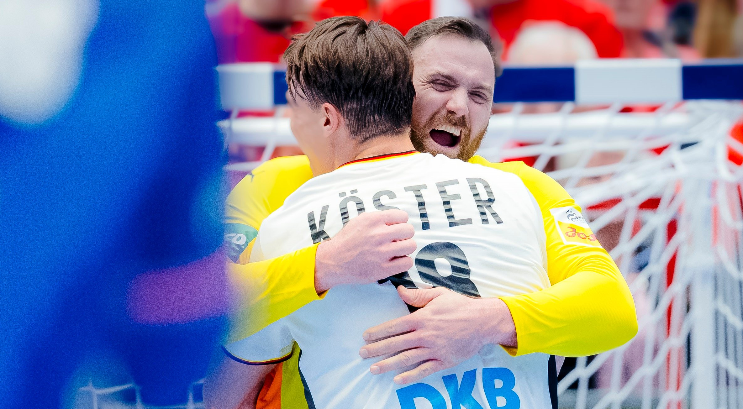 Zwei männliche Handballspieler in weißen Trikots umarmen sich und feiern fröhlich auf dem Spielfeld vor einem Tornetz, wobei der Name Köster" und die Nummer 19 auf dem Rücken des einen Spielers zu sehen sind.