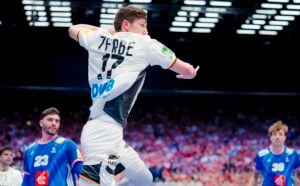 Ein Handballspieler in einem weißen Trikot springt zum Wurf, mit der Zerbe 17 auf dem Rücken. Zwei gegnerische Teammitglieder in Blau schauen ihm in einer hell erleuchteten Halle zu.