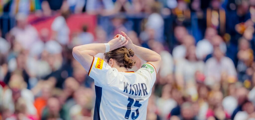 Ein Handballspieler, der ein weißes Trikot mit dem Namen KNORR und der Nummer 15 trägt, steht mit über dem Kopf zusammengeschlagenen Händen, von der Kamera abgewandt, vor einem verschwommenen, überfüllten Publikum.