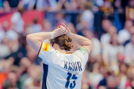 Ein Handballspieler, der ein weißes Trikot mit dem Namen KNORR und der Nummer 15 trägt, steht mit über dem Kopf zusammengeschlagenen Händen, von der Kamera abgewandt, vor einem verschwommenen, überfüllten Publikum.