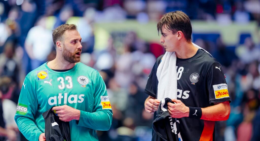 Zwei männliche Handballspieler in schwarzen und blaugrünen Trikots stehen nebeneinander, halten Handtücher und unterhalten sich während eines Spiels, mit einer unscharfen Menschenmenge im Hintergrund.