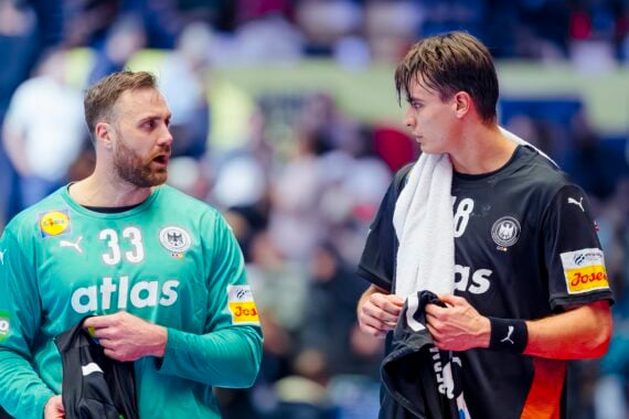 Zwei männliche Handballspieler in schwarzen und blaugrünen Trikots stehen nebeneinander, halten Handtücher und unterhalten sich während eines Spiels, mit einer unscharfen Menschenmenge im Hintergrund.