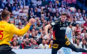 Ein männlicher Handballspieler in einem schwarzen Trikot bereitet sich darauf vor, den Ball zu werfen, während er einem gelb gekleideten Torwart gegenübersteht. Das Spiel findet in einer überfüllten Arena statt, die von Zuschauern aufmerksam verfolgt wird.