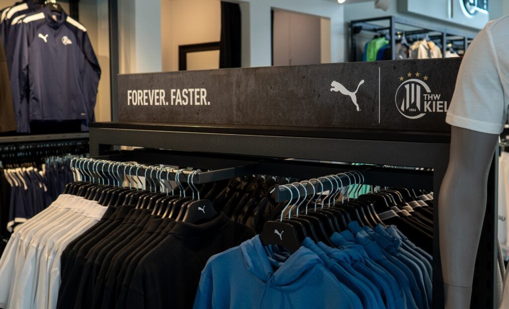 Auf den Kleiderständern in einem Geschäft ist Puma-Sportbekleidung mit schwarzen, blauen und weißen Hemden und Kapuzenpullis ausgestellt. Auf einem Schild darüber steht FOREVER. FASTER. neben dem Puma-Logo und dem Mannschaftsemblem des THW Kiel.