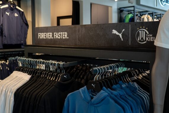 Auf den Kleiderständern in einem Geschäft ist Puma-Sportbekleidung mit schwarzen, blauen und weißen Hemden und Kapuzenpullis ausgestellt. Auf einem Schild darüber steht FOREVER. FASTER. neben dem Puma-Logo und dem Mannschaftsemblem des THW Kiel.