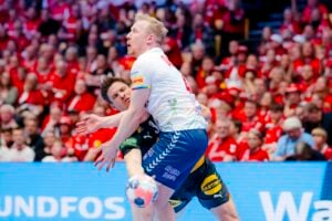 Zwei männliche Handballspieler in Aktion während eines Spiels, der eine in einem weiß-blauen und der andere in einem schwarzen Trikot, umgeben von einer überwiegend in Rot gekleideten Zuschauermenge.