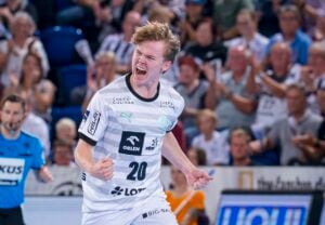 Ein männlicher Handballspieler in einem weiß-grauen Trikot mit der Nummer 20 ballt die Fäuste und jubelt auf dem Spielfeld, während die Fans im Hintergrund jubeln. In der Nähe ist auch ein Offizieller in Blau zu sehen.