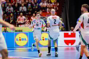 Zwei männliche Handballspieler in weißen Trikots, einer mit der Nummer 91 und der andere mit der Nummer 2, laufen während eines Spiels auf einem Hallenplatz. Eine Menschenmenge schaut von der Tribüne im Hintergrund zu.