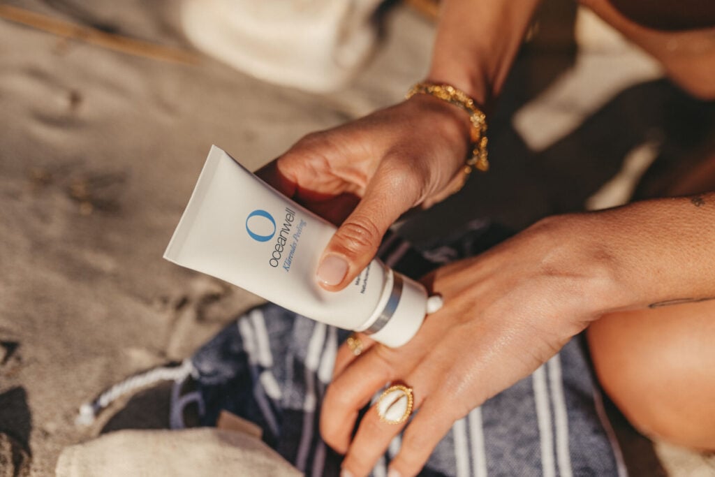 Am Strand sitzend, mit Goldschmuck und einer gestreiften Stofftasche in der Hand, cremt sich eine Person den Handrücken mit Oceanwell ein - und unterstützt mit jeder wohltuenden Berührung das THW Kiel För De Küste Nachhaltigkeit Projekt.