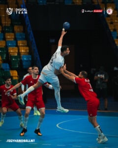 Ein Handballspieler in einem weißen Trikot springt hoch, um den Ball zu schießen, während ein Verteidiger in einem roten Trikot versucht, ihn zu blockieren. Im Hintergrund sind andere Spieler und leere Sitzreihen zu sehen.