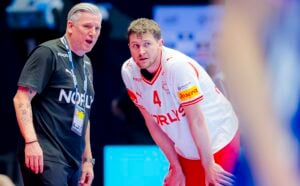 Ein Volleyballtrainer und ein Spieler in NORLYS-Trikots unterhalten sich während eines Spiels. Der Trainer scheint Anweisungen zu geben, während der Spieler aufmerksam zuhört, die Hände auf den Knien.